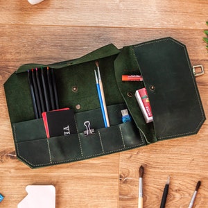 May include: A dark green leather pencil case with multiple pockets and compartments. The case is open and filled with pencils, pens, brushes, and other art supplies.