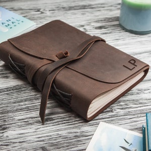 May include: A brown leather journal with a strap closure and the initials "LP" embossed on the cover. The journal is lying on a wooden surface.
