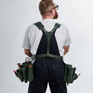 Puede incluir: Una persona que lleva una camiseta blanca y un mono de trabajo de cuero verde oscuro con un cinturón de herramientas. El cinturón de herramientas tiene varios bolsillos y está lleno de herramientas.