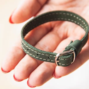 May include: A green leather bracelet with white stitching and a silver buckle.