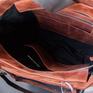 K&ouml;nnte beinhalten: Das Innere einer braunen Ledertasche mit schwarzem Stofffutter. Die Tasche hat eine Rei&szlig;verschlusstasche und eine kleinere offene Tasche im Inneren.