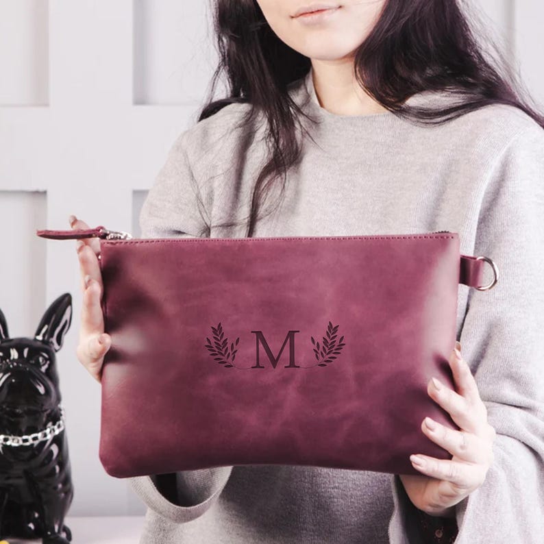 Peut inclure: Une pochette en cuir bordeaux avec une fermeture &eacute;clair et une sangle argent&eacute;e. La pochette est monogramme avec l'initiale "M" et un motif de feuilles.