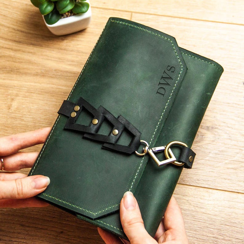 May include: A dark green leather journal with a black strap and metal accents. The journal has the initials "DWS" embossed on the front.