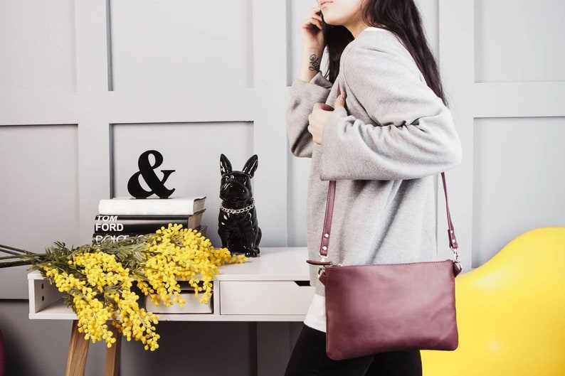 Peut inclure: Un sac &agrave; bandouli&egrave;re en cuir bordeaux avec une fermeture &eacute;clair argent&eacute;e et une longue bandouli&egrave;re. Le sac est port&eacute; par une personne portant un pull gris.
