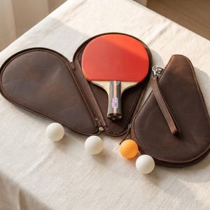 May include: A ping pong paddle with a red rubber surface rests in a brown leather case. Three white and one orange ping pong balls are arranged on a white surface. The paddle handle has a black and wood grain design.