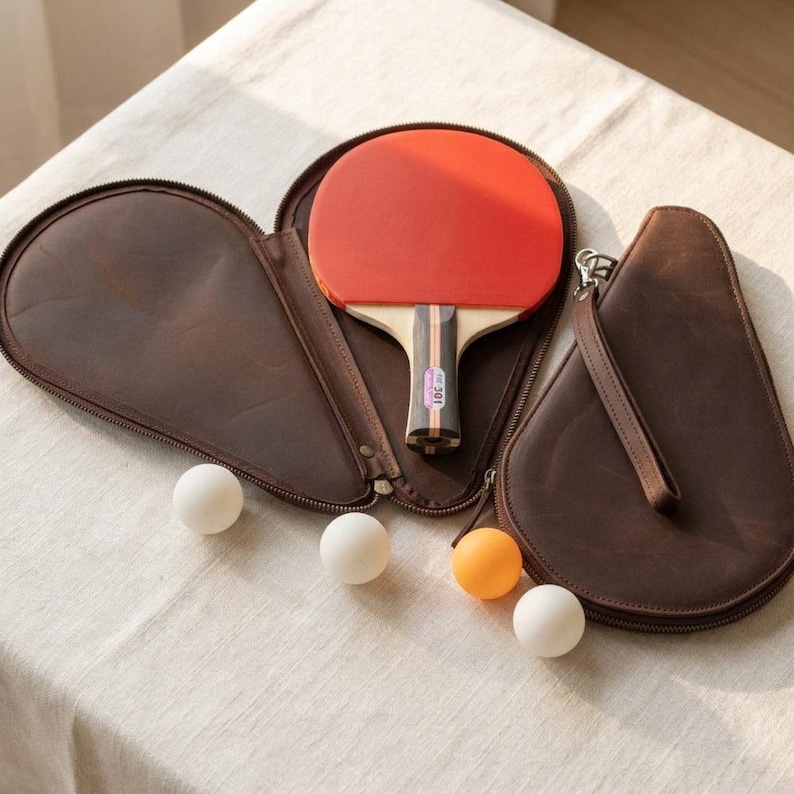 May include: A ping pong paddle with a red rubber surface rests in a brown leather case. Three white and one orange ping pong balls are arranged on a white surface. The paddle handle has a black and wood grain design.