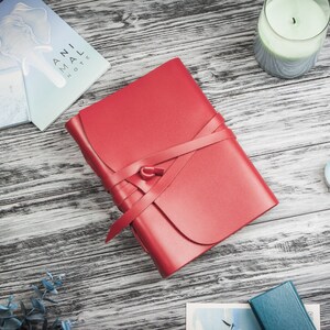 May include: A red leather journal with a ribbon closure. The journal is on a wooden surface with other items, including a candle and a small piece of paper.