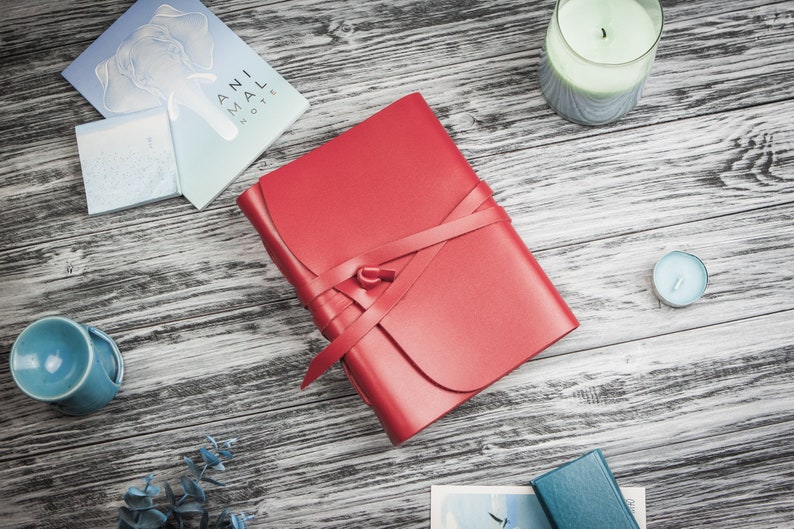 May include: A red leather journal with a ribbon closure. The journal is on a wooden surface with other items, including a candle and a small piece of paper.