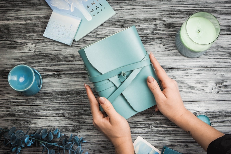 May include: A teal leather journal with a criss-cross strap closure. The journal is being held by a person's hands. The journal is on a wooden surface with a candle and other items.