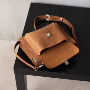 May include: A brown leather satchel bag with a flap closure and a silver buckle. The bag has a long strap and a smaller pocket on the front.