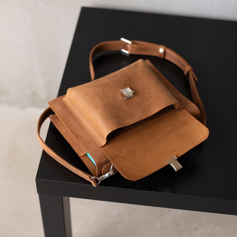 May include: A brown leather satchel bag with a flap closure and a silver buckle. The bag has a long strap and a smaller pocket on the front.