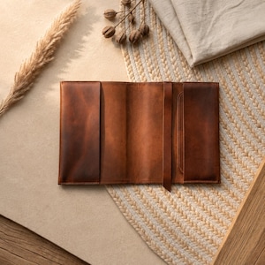 May include: A rectangular brown leather cover featuring a smooth, matte finish with two side flaps and a central vertical strap. The leather displays natural tonal variations in a rich, warm brown hue, designed to fold over and protect a book or journal.