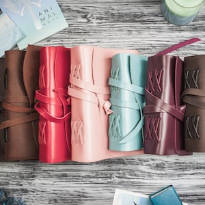 May include: Seven leather-bound journals in different colors, including brown, red, pink, blue, burgundy, and brown. Each journal has a leather strap and decorative stitching.