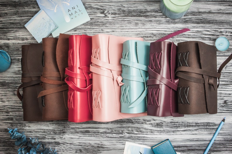May include: Seven leather-bound journals in different colors, including brown, red, pink, blue, burgundy, and brown. Each journal has a leather strap and decorative stitching.