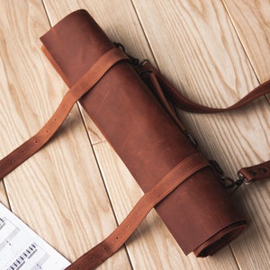 May include: A brown leather roll-up case with a long strap. The case is rolled up and secured with a strap. The case is on a wooden surface.