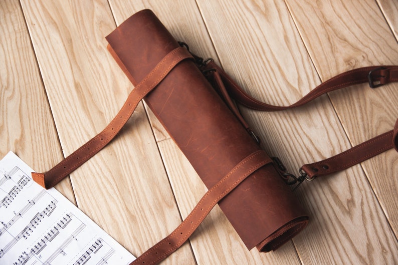 May include: A brown leather roll-up case with a long strap. The case is rolled up and secured with a strap. The case is on a wooden surface.
