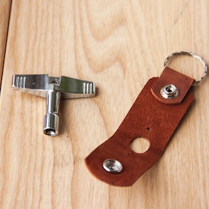 May include: A silver drum key and a brown leather strap with a metal ring. The strap has a hole in the center and a metal button on the top.
