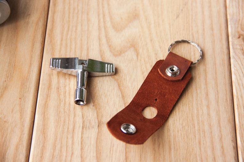 May include: A silver drum key and a brown leather strap with a metal ring. The strap has a hole in the center and a metal button on the top.