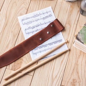 May include: A brown leather drumstick holder with a sheet of music and two wooden drumsticks on a wooden surface.
