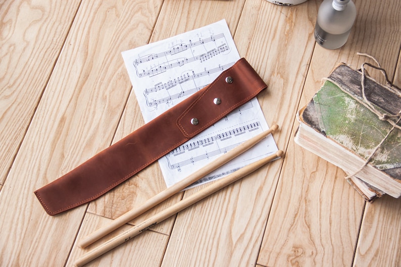 May include: A brown leather drumstick holder with a sheet of music and two wooden drumsticks on a wooden surface.