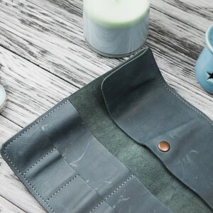 May include: A teal leather travel case with multiple slots for holding pens, pencils, or other writing utensils. The case is open and lying flat on a light wood surface.