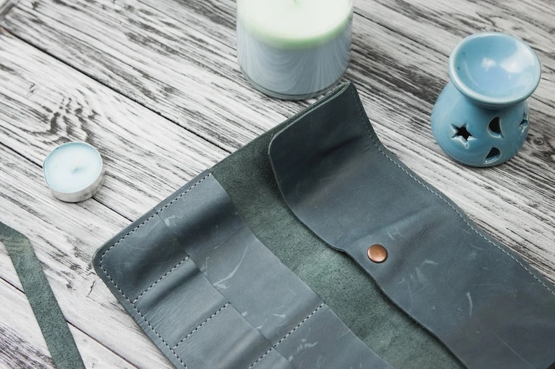 May include: A teal leather travel case with multiple slots for holding pens, pencils, or other writing utensils. The case is open and lying flat on a light wood surface.