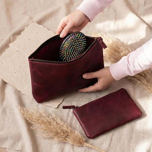Pu&ograve; includere: Un beauty case in pelle bordeaux e una piccola pochette abbinata. All'interno della borsa aperta c'&egrave; una spazzola per capelli nera con setole colorate. Le borse sono su una superficie neutra.