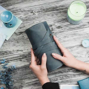 May include: A blue leather notebook with a strap closure, held by a person's hands on a wooden surface.