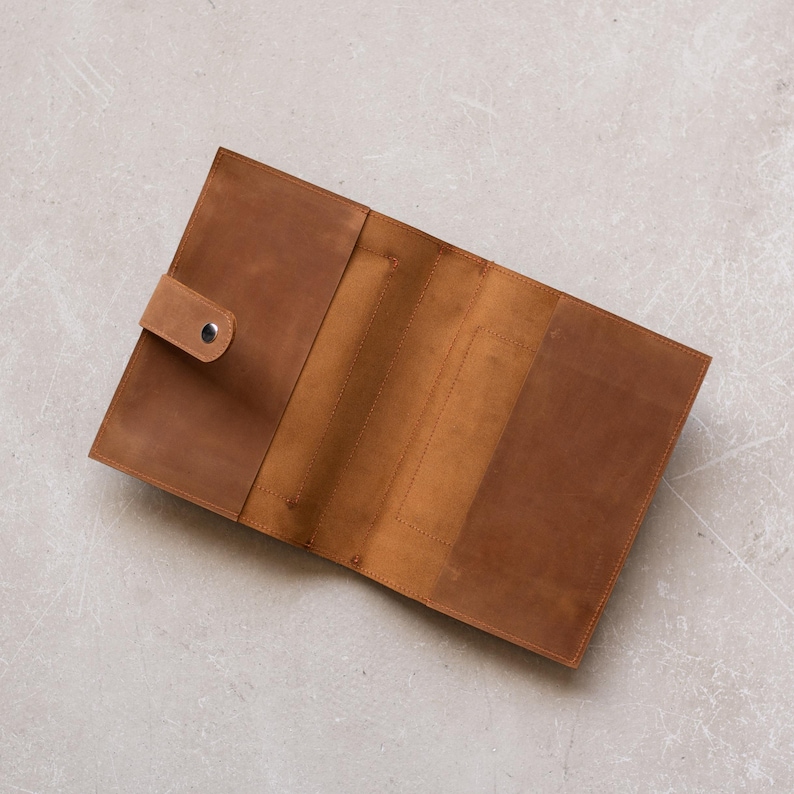 May include: A brown leather notebook cover with a flap closure and a button. The cover is open to reveal the interior, which has multiple pockets and slots for organization.