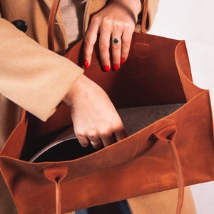 May include: A brown leather tote bag with a gray fabric lining. The bag is open and a person is placing a black item inside.