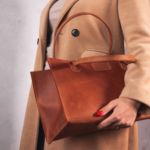 May include: A brown leather tote bag with a top handle. The bag is held by a person wearing a beige coat.