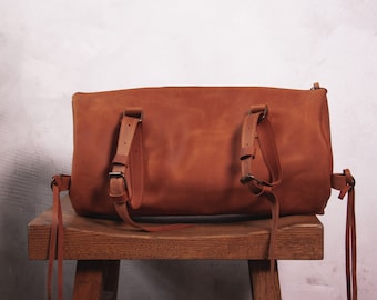 Puede incluir: Una mochila de cuero marrón con correas ajustables y lazos largos de cuero. La mochila está sentada en un taburete de madera.