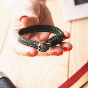 May include: A dark green leather camera strap with a silver buckle. The strap is stitched with white thread.