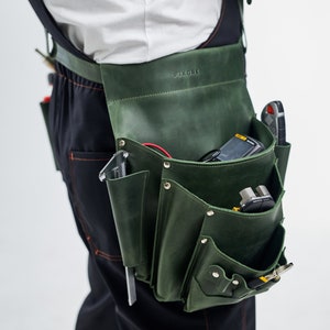 K&ouml;nnte beinhalten: Ein gr&uuml;ner Leder-Werkzeugg&uuml;rtel mit mehreren Taschen und F&auml;chern, getragen &uuml;ber einer schwarzen Arbeitshose. Der G&uuml;rtel ist mit "PIKORE" beschriftet und mit verschiedenen Werkzeugen gef&uuml;llt, darunter ein Multimeter, Drahtschneider und ein Schraubendreher.