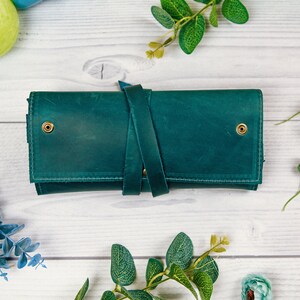 May include: A teal leather wallet with a strap closure and brass accents. The wallet is on a white wooden surface.