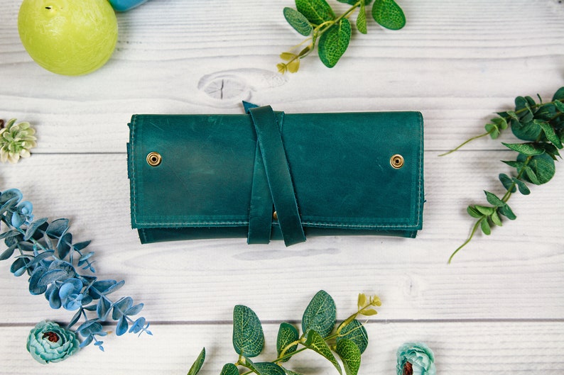 May include: A teal leather wallet with a strap closure and brass accents. The wallet is on a white wooden surface.