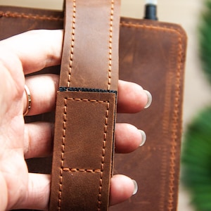 May include: A brown leather notebook cover with a brown leather strap and black stitching. The strap has a black elastic band.