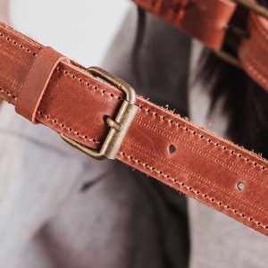 May include: Close-up of a brown leather strap with a brass buckle. The strap has stitching along the edges and is designed for a bag or camera.