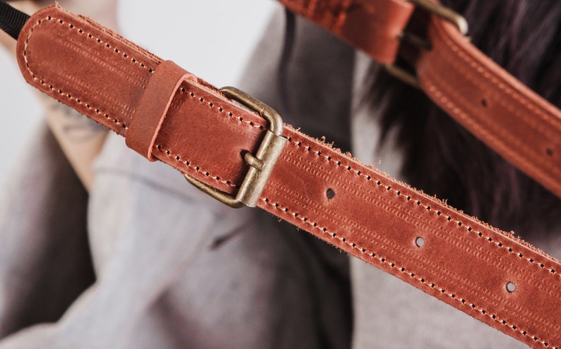May include: Close-up of a brown leather strap with a brass buckle. The strap has stitching along the edges and is designed for a bag or camera.