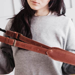 May include: A brown leather camera strap with two adjustable buckles. The strap is shown being held by a person.