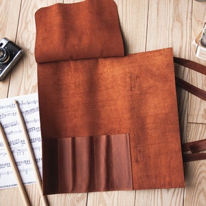 May include: A brown leather roll-up case with multiple pockets for storing drumsticks. The case is laid out flat on a wooden surface with sheet music and drumsticks beside it.