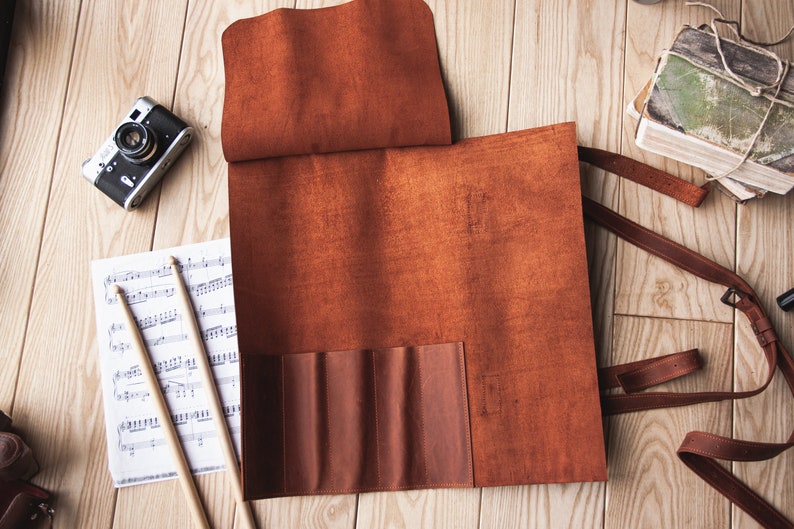 May include: A brown leather roll-up case with multiple pockets for storing drumsticks. The case is laid out flat on a wooden surface with sheet music and drumsticks beside it.