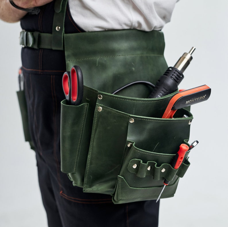 May include: A green leather tool belt with multiple pockets and compartments. The belt is worn by a person and contains various tools, including a pair of scissors, a soldering iron, and a screwdriver.