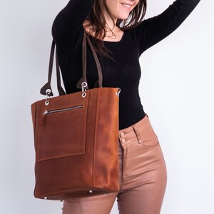 K&ouml;nnte beinhalten: Eine braune Ledertasche mit einer Rei&szlig;verschlusstasche auf der Vorderseite. Die Tasche hat einen Rei&szlig;verschluss oben und zwei obere Tragegriffe.