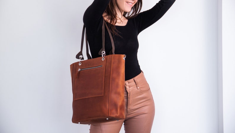 K&ouml;nnte beinhalten: Eine braune Ledertasche mit einer Rei&szlig;verschlusstasche auf der Vorderseite. Die Tasche hat einen Rei&szlig;verschluss oben und zwei obere Tragegriffe.
