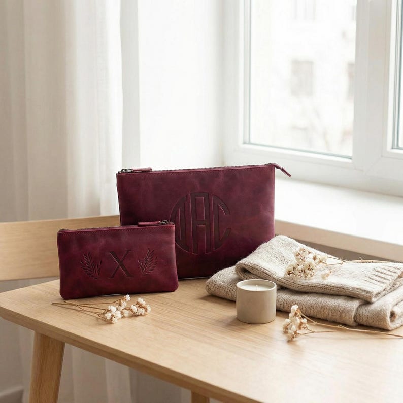 Pu&ograve; includere: Pochette in pelle bordeaux con chiusure a zip. La pochette pi&ugrave; grande presenta un monogramma, mentre quella pi&ugrave; piccola mostra una "X" con accenti di foglie. Una coperta piegata, una candela e fiori secchi sono su un tavolo di legno.