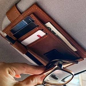 May include: Brown leather car visor organizer with multiple card slots and a pen holder. A pair of glasses is hanging from the organizer.