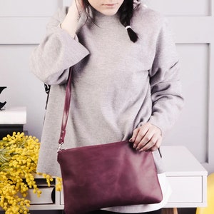 Peut inclure: Une femme portant un pull gris et un sac bandouli&egrave;re bordeaux. Le sac a une longue bandouli&egrave;re et une fermeture &agrave; rabat. La femme est debout devant un mur blanc avec un signe ampersand noir sur une &eacute;tag&egrave;re.