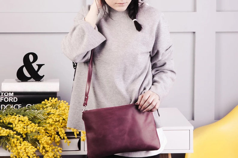 Peut inclure: Une femme portant un pull gris et un sac bandouli&egrave;re bordeaux. Le sac a une longue bandouli&egrave;re et une fermeture &agrave; rabat. La femme est debout devant un mur blanc avec un signe ampersand noir sur une &eacute;tag&egrave;re.