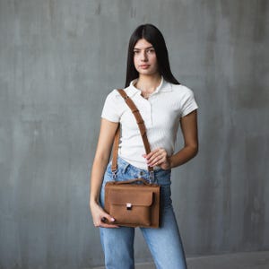 May include: A woman wearing a white collared shirt and blue jeans is carrying a brown leather satchel bag with a shoulder strap.